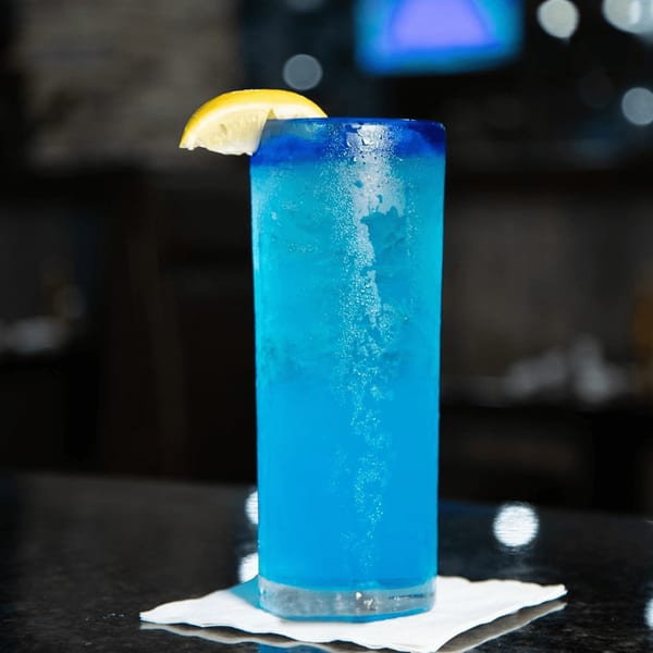 A vibrant blue cocktail with a lemon wedge, condensation on the glass, on a dark bar.