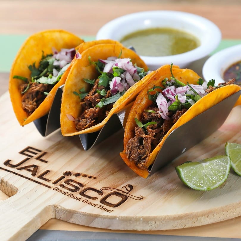 Three birria tacos with red onions and cilantro, served with lime and two sauces on a wooden board.