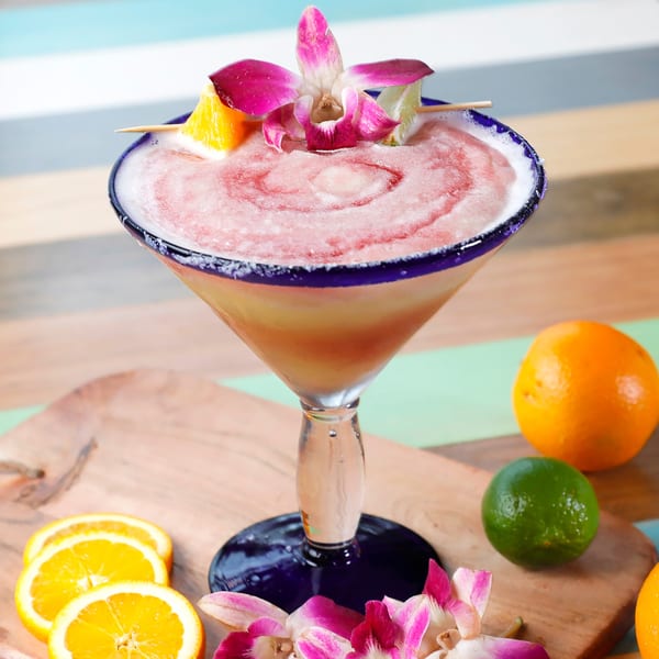 Pink frozen cocktail in a blue-rimmed glass, garnished with an orchid, orange, and lime.
