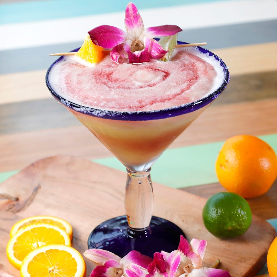 Pink frozen cocktail in a blue-rimmed glass, garnished with an orchid, orange, and lime.