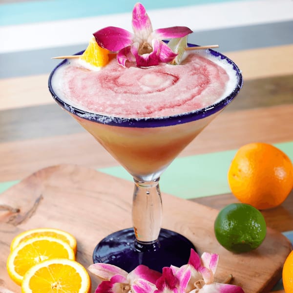 Pink frozen margarita in a blue-rimmed glass, garnished with an orchid, orange, and lime.